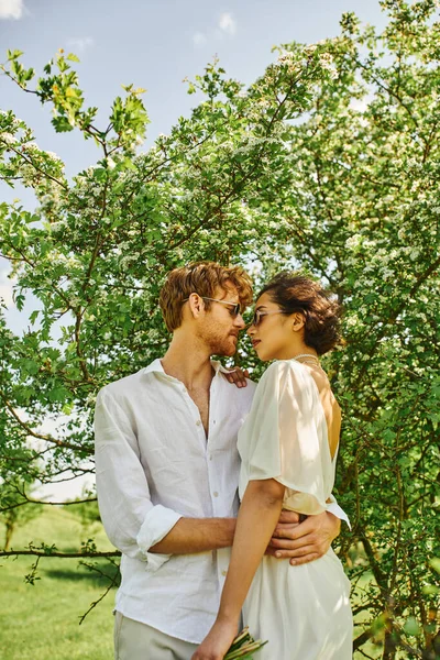Felices recién casados multiétnicos en gafas de sol y vestido de novia abrazándose en el campo, estilo boho - foto de stock