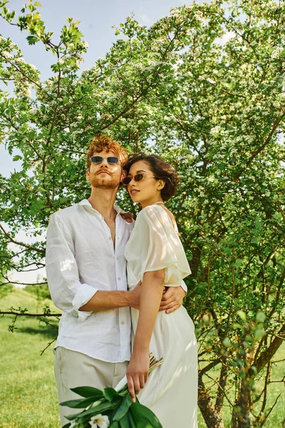 Щасливі багатоетнічні молодята в сонцезахисних окулярах і весільних сукнях обіймаються в зеленому саду, стилі бохо — стокове фото