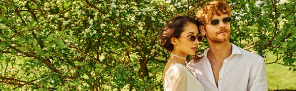 Молоді багатоетнічні молодята в сонцезахисних окулярах і весільній сукні обіймаються в зеленому саду, банер — стокове фото