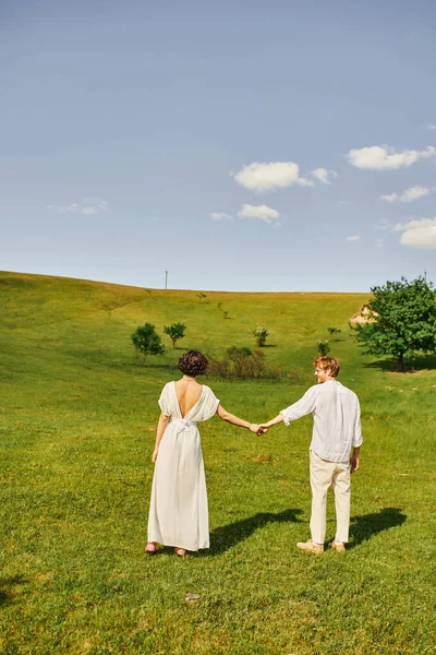 Happy redhead man holding hands with bride in white dress and standing together in green field — Stock Photo
