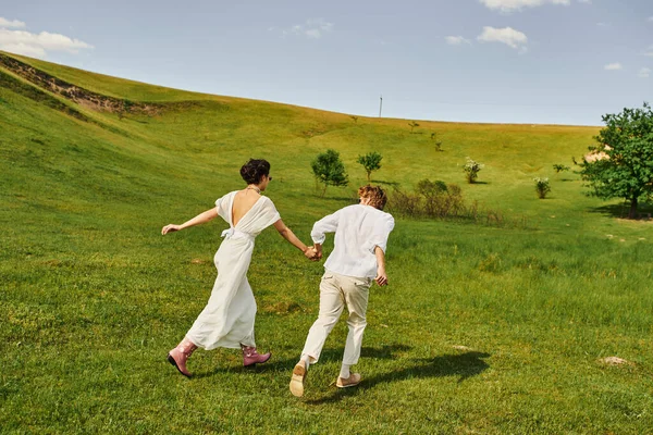 Vista trasera de un par de recién casados corriendo en campo verde, boda rústica, atuendo blanco estilo boho - foto de stock