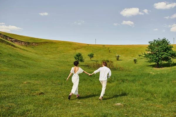 Young couple of newlyweds running in green field, bride and groom in wedding gown holding hands — Stock Photo