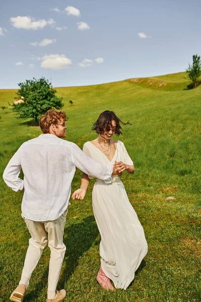 Feliz recién casados sonriendo y bailando en verde campo, asiático novia en vestido de novia y pelirroja novio - foto de stock