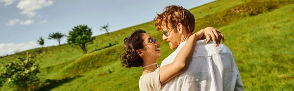 Feliz recién casados sonriendo y abrazándose en verde campo, novia asiática en vestido blanco y novio, bandera - foto de stock