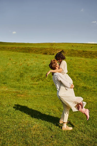 Alegre casal, feliz asiático mulher no branco vestido e cowboy botas abraçando noivo no verde campo — Fotografia de Stock