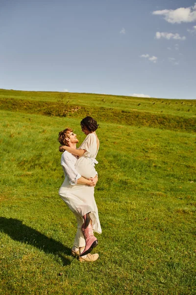 Sposi gioiosi, sposo felice sollevamento donna asiatica in abito bianco e stivali cowboy in campo verde — Foto stock