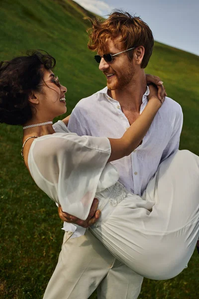 Щасливі молодята, радісний наречений, що піднімає азіатську наречену в білій сукні в зеленому полі, сільська природа — стокове фото