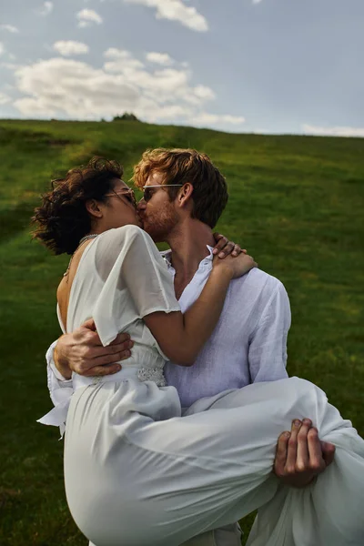Sposi felici, sposo sollevamento e baci sposa asiatica in abito bianco in campo verde, campagna — Foto stock