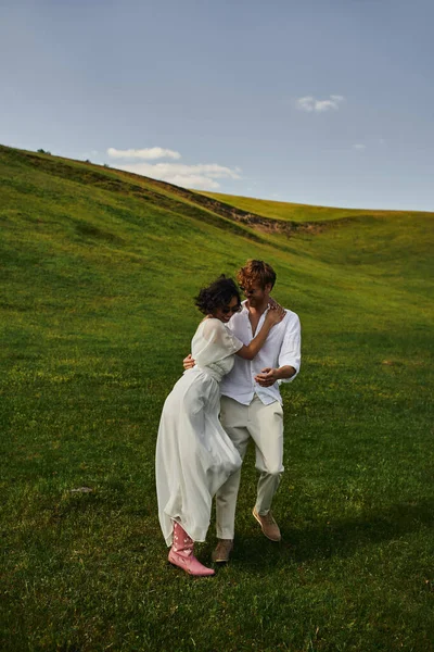 Happy newlyweds, asian bride in white dress and cowboy boots walking with groom in green field — Stock Photo