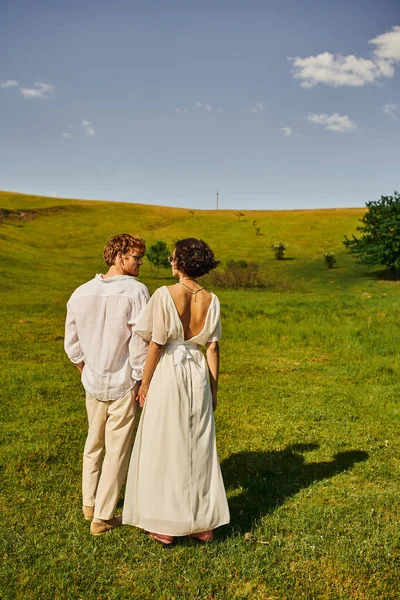 Just married couple, asian bride in wedding dress holding hands with redhead groom in green field — Stock Photo