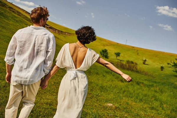 Matrimonio rurale, vista posteriore, sposa in abito da sposa che cammina con lo sposo in campo, coppia appena sposata — Foto stock