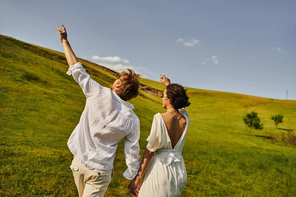 Rural wedding, just married couple in wedding gown holding hands and walking in green field — Stock Photo