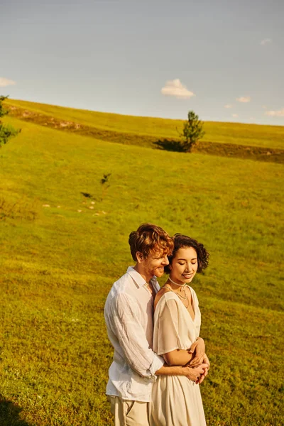 Just married couple, happy man hugging asian bride in green field, scenic and tranquil landscape — Stock Photo