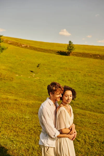 Pareja recién casada, hombre feliz abrazando novia asiática en el campo verde, paisaje escénico y tranquilo - foto de stock