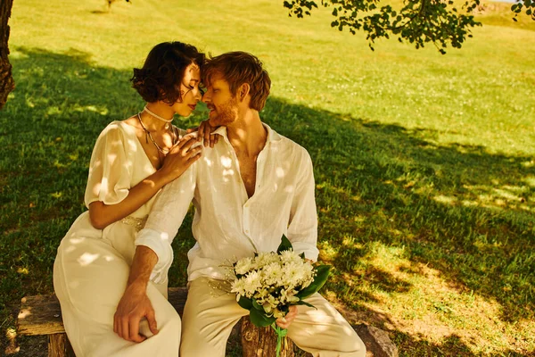 Boda rural, interracial pareja recién casada sentado bajo el árbol con ramo de novia, recién casados - foto de stock