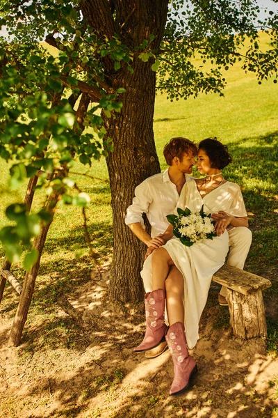 Redhead groom embracing asian bride with bouquet while sitting on bench under tree, rustic wedding — Stock Photo