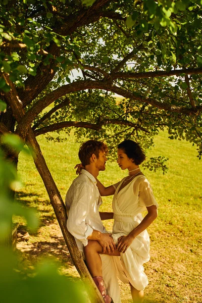 Side view of interracial boho style couple near ladder and tree in countryside, rural wedding — Stock Photo