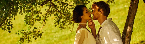 Marié rousse embrasser charmante mariée asiatique sous arbre vert dans un cadre rural, vue de côté, bannière — Photo de stock