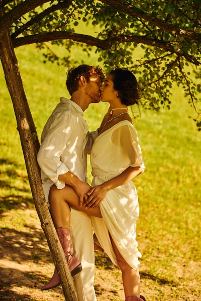 Interracial newlyweds in boho style wedding attire kissing under tree near ladder, rural setting — Stock Photo