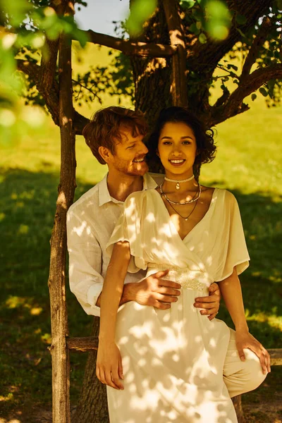 Mariage rustique, souriant rousse marié étreignant joyeuse mariée asiatique sous l'arbre le jour ensoleillé — Photo de stock