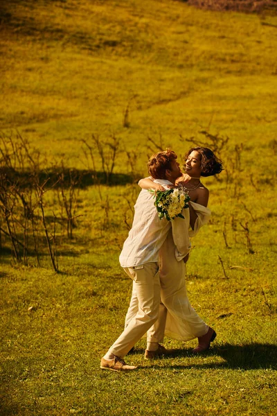 Marié rousse embrassant joyeuse mariée asiatique avec des fleurs dans le champ vert, célébration rurale — Photo de stock