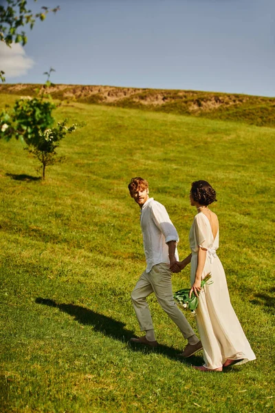 Pareja interracial recién casada en traje de boda blanco tomados de la mano y corriendo en el prado verde - foto de stock
