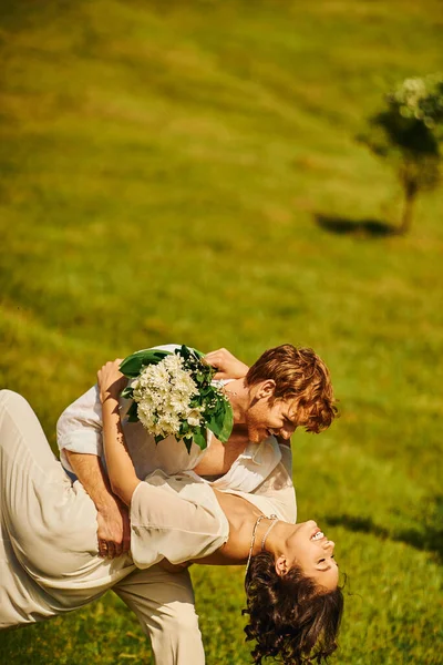 Marié rousse embrassant heureuse mariée asiatique avec des fleurs sur prairie verte, mariage rustique, vue latérale — Photo de stock