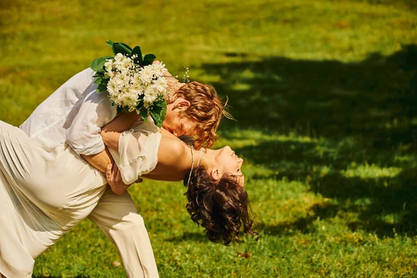 Young redhead groom hugging and kissing asian bride with flowers on green meadow, rustic wedding — Stock Photo