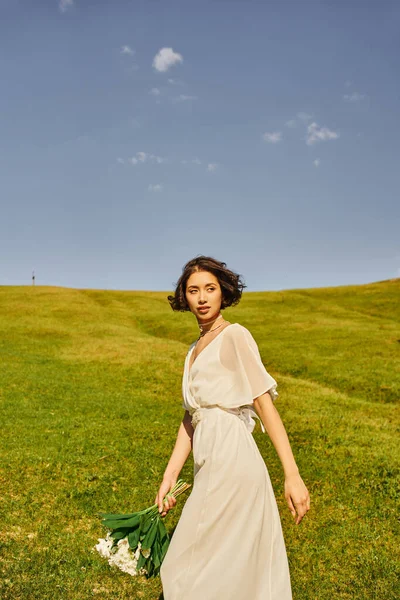 Encantadora noiva asiática no estilo boho vestido andando com buquê de casamento no prado verde, ambiente rural — Fotografia de Stock