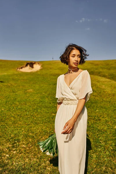 Брюнетка азіатська жінка в стилі бохо весільна сукня тримає квіти і дивиться в зелене поле — стокове фото