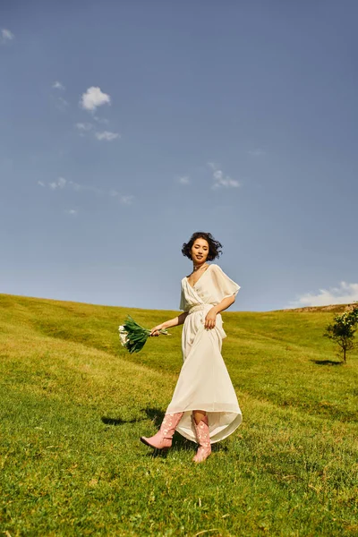 Carefree asian woman in wedding dress and cowboy boots walking with flowers in green field — Stock Photo