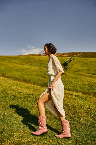 Side view of young asian woman in bridal dress and cowboy boots walking on meadow in countryside — Stock Photo