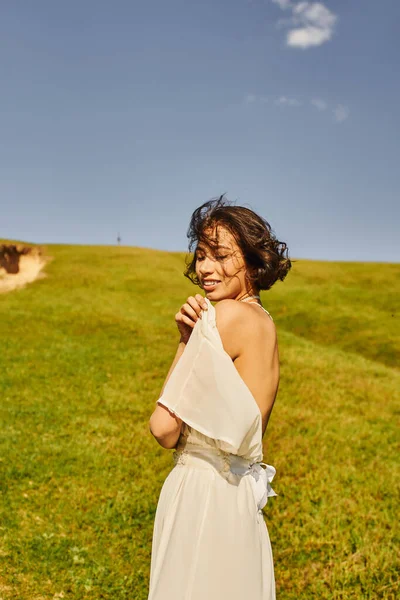 Sensual asian bride in boho style dress smiling on green meadow under blue sky, serenity and joy — Stock Photo