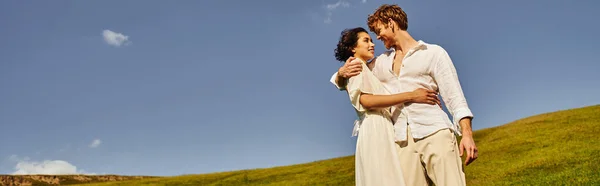 Felices recién casados multiétnicos abrazándose en el prado verde bajo el cielo azul, boda en un entorno rural, pancarta - foto de stock