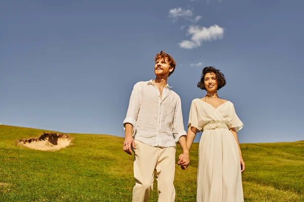 Jovem, despreocupado inter-racial recém-casados de mãos dadas no campo verde sob o céu azul, paisagem cênica — Fotografia de Stock