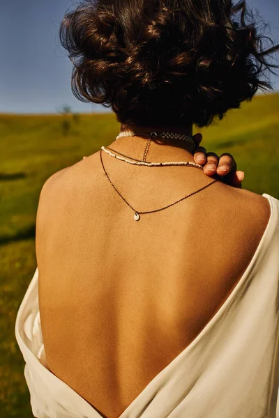 Youthful woman in necklaces and bridal gown with naked back in countryside, serenity and happiness — Stock Photo