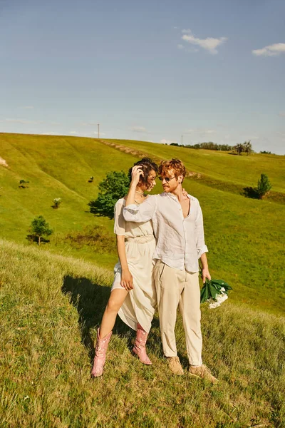 Jóvenes interracial recién casados en traje de estilo boho en pradera verde, unión y felicidad en el campo - foto de stock