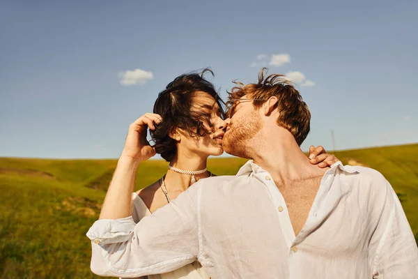 Frisch verheiratete gemischtrassige Paare küssen sich bei Wind unter blauem Himmel, rustikale Hochzeit in ruhiger Umgebung — Stockfoto