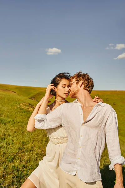 Multiethnic newlywed couple in white wedding attire kissing on green meadow, serenity and tenderness — Stock Photo