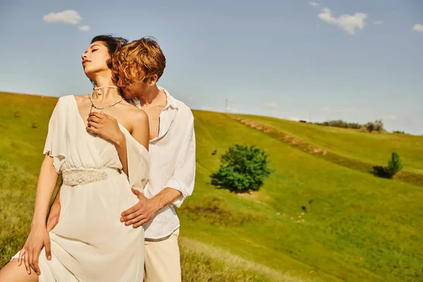 Ruiva homem em óculos de sol abraçando elegante noiva asiática no prado verde, tranquilo casamento rústico — Fotografia de Stock