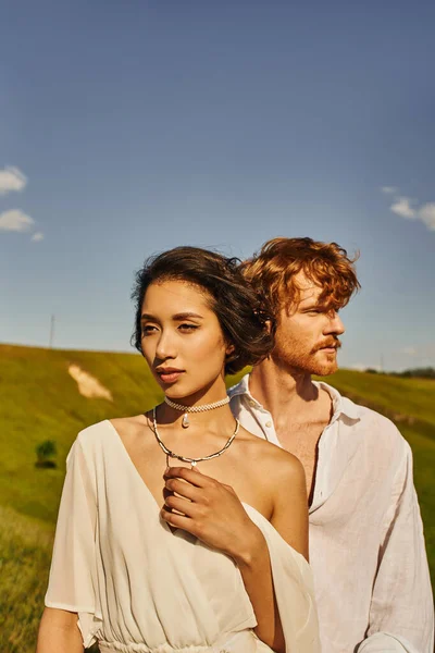 Charmante femme asiatique dans le style boho robe de mariée près de jeune marié rousse dans un cadre rural pittoresque — Photo de stock