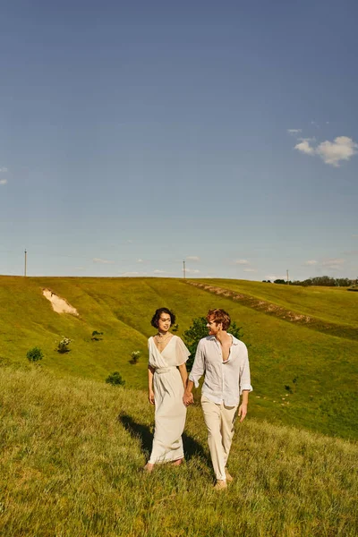 Rural romance, stylish multiethnic newlyweds holding hands and walking on green field under blue sky — Stock Photo