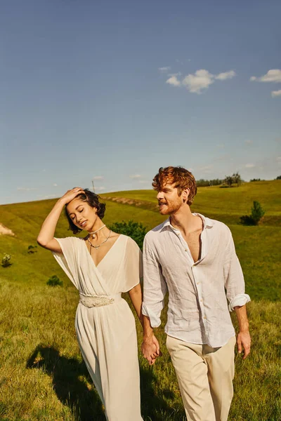 Romantisches gemischtrassiges Brautpaar Händchen haltend und auf dem Land mit malerischer Landschaft spazierend — Stockfoto