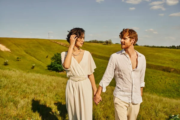Pareja interracial recién casada tomados de la mano y sonriéndose en el prado, el amor y la serenidad - foto de stock