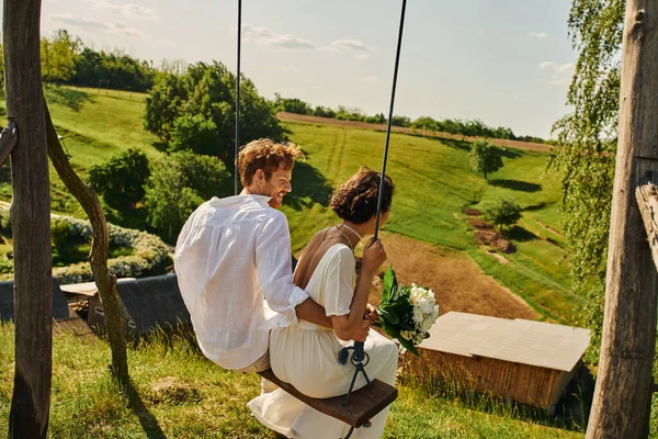 Cheerful redhead groom swinging with bride in white dress in countryside with picturesque landscape — Stock Photo