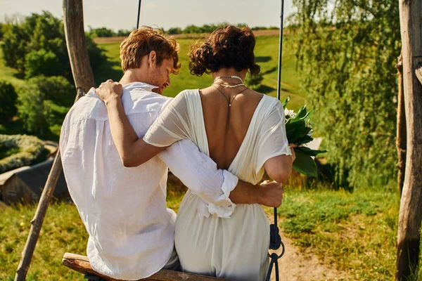 Back view of stylish just married couple embracing on swing in countryside, rustic wedding — Stock Photo