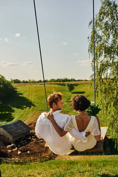 Vue arrière des jeunes mariés sur le swing à la campagne avec ferme et collines verdoyantes, amour et sérénité — Photo de stock