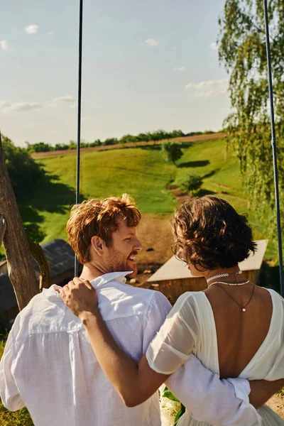 Young interracial couple on swing in picturesque countryside setting, rural wedding — Stock Photo