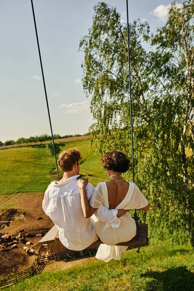 Rustic wedding, back view of redhead groom with young bride swinging in picturesque countryside — Stock Photo