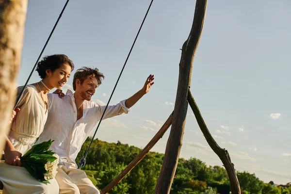 Joyful redhead groom waving hand near asian bride with flowers on swing in scenic rustic setting — Stock Photo